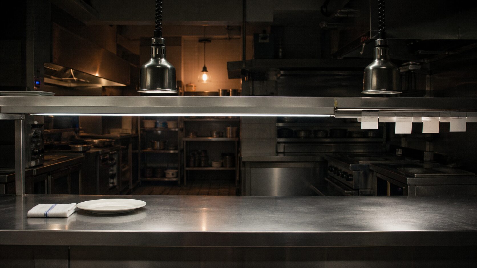 A luxury restaurant kitchen pass after service has ended, stainless steel counter empty, heat lamps off, a single plate and folded service towel